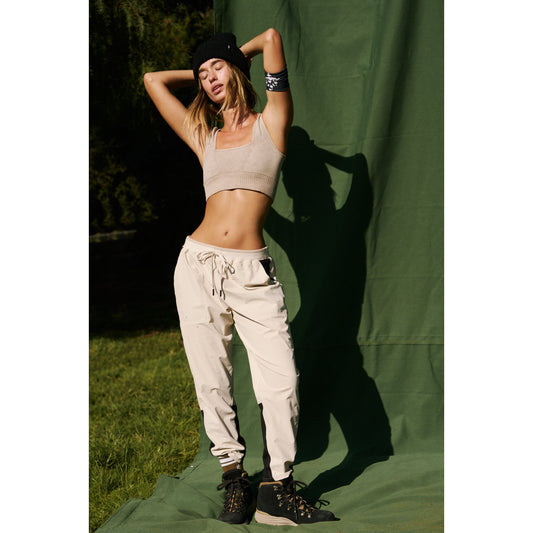 A woman in Free People Movement's Cascade Jogger in Muted Beige stands against a green backdrop, posing with her hands behind her head.