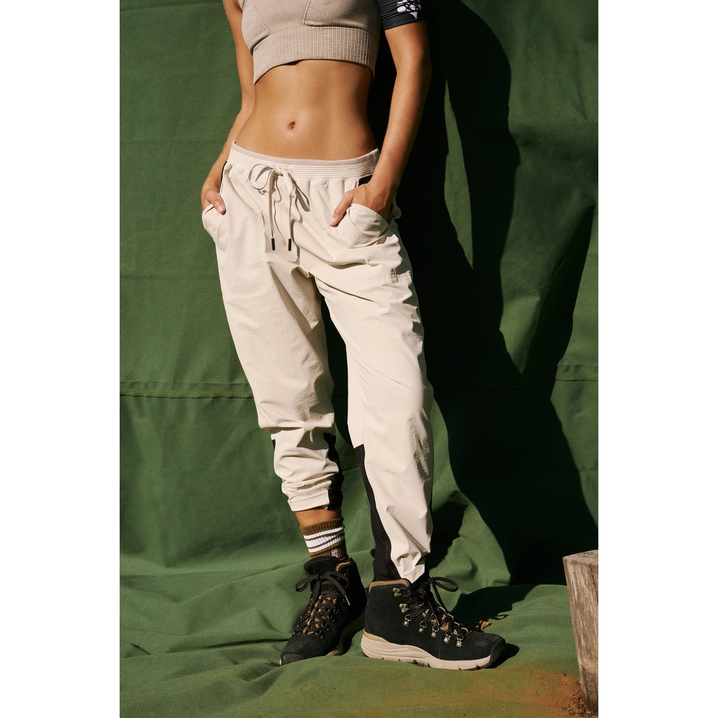 A person wearing a light grey crop top and high-waisted Muted Beige Cascade Joggers stands with hands in pockets against a green backdrop, only the torso and legs are visible. (Brand: Free People Movement)