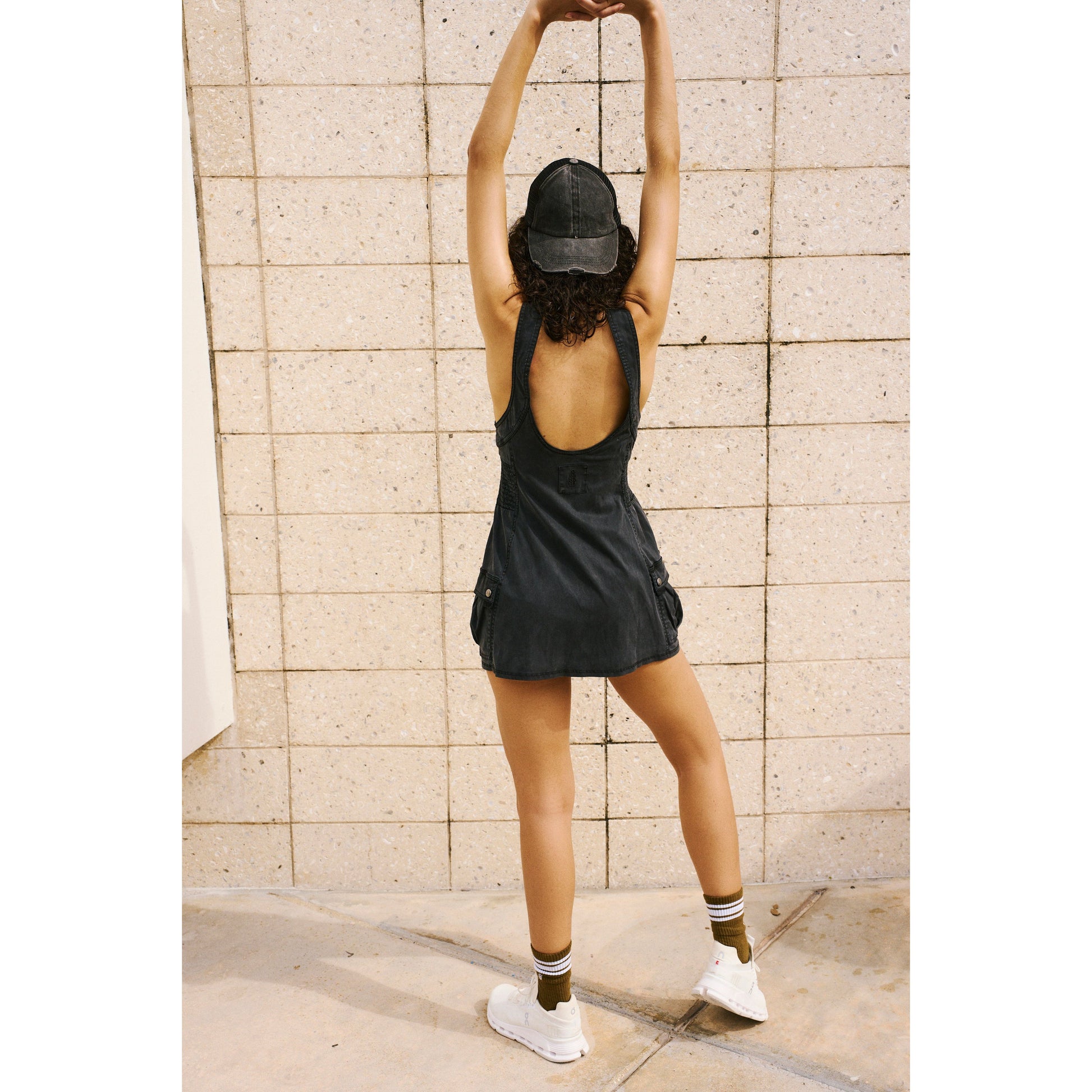 A woman in a dark Fast Track Skortsie by Free People Movement with cargo-inspired fabrication and cap stretches her arms upwards against a beige tiled wall, wearing white sneakers and striped socks.