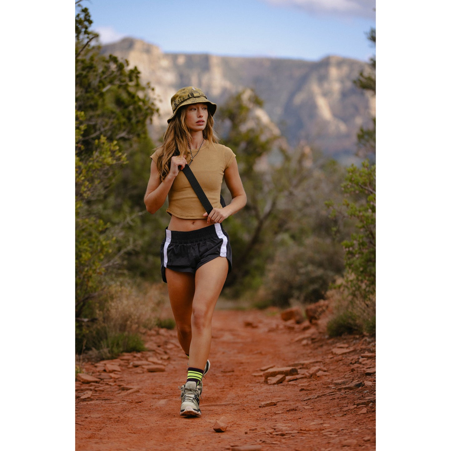 A woman in Free People Movement's Trail Angel Shorts in Black Combo and hiking gear walks on a dirt trail with mountains in the background.