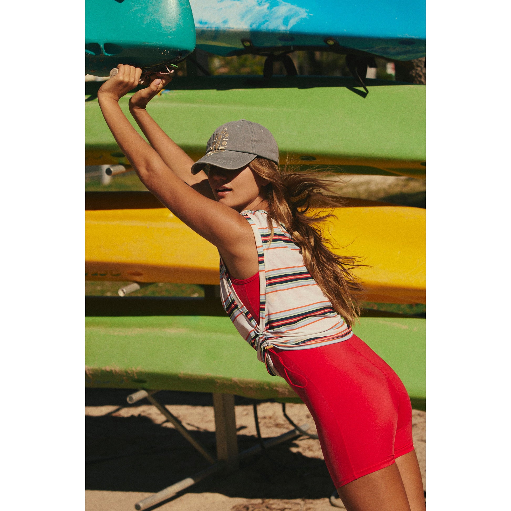 A woman in a red skirt and striped top lifts a blue kayak onto a rack with colorful kayaks in bright sunlight wearing the Sport Mode Muscle Tee in Electric Combo by Free People Movement.