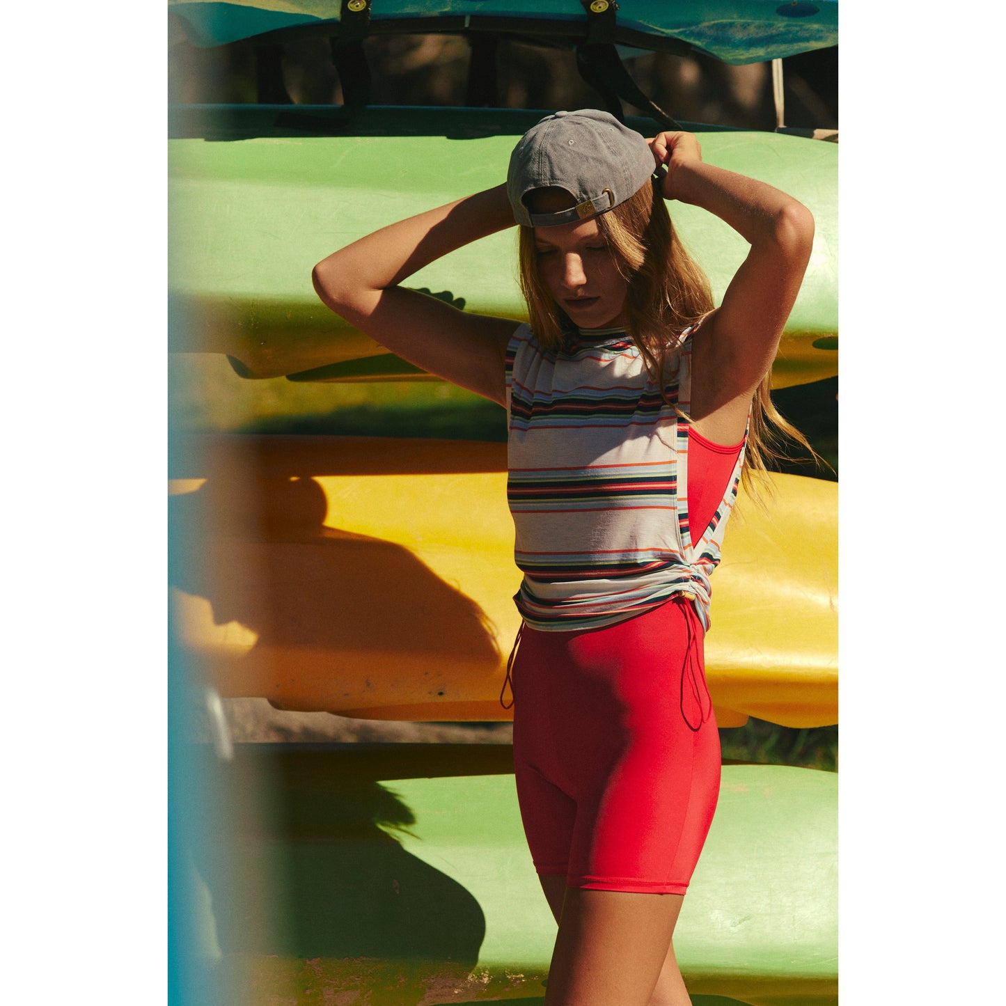 A woman in a Free People Movement Sport Mode Muscle Tee and Electric Combo shorts adjusts her gray cap near a stack of colorful kayaks in sunlight.