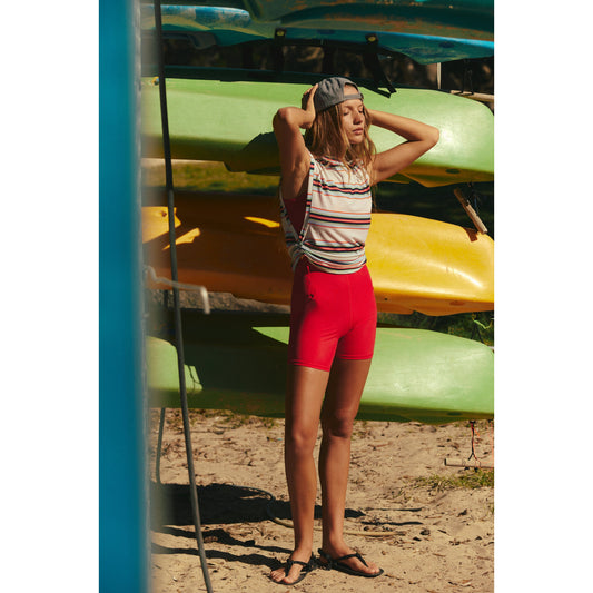 Young woman in a Sport Mode Muscle Tee by Free People Movement and red shorts standing by colorful kayaks, adjusting a gray cap on her head, outdoors on a sunny day.