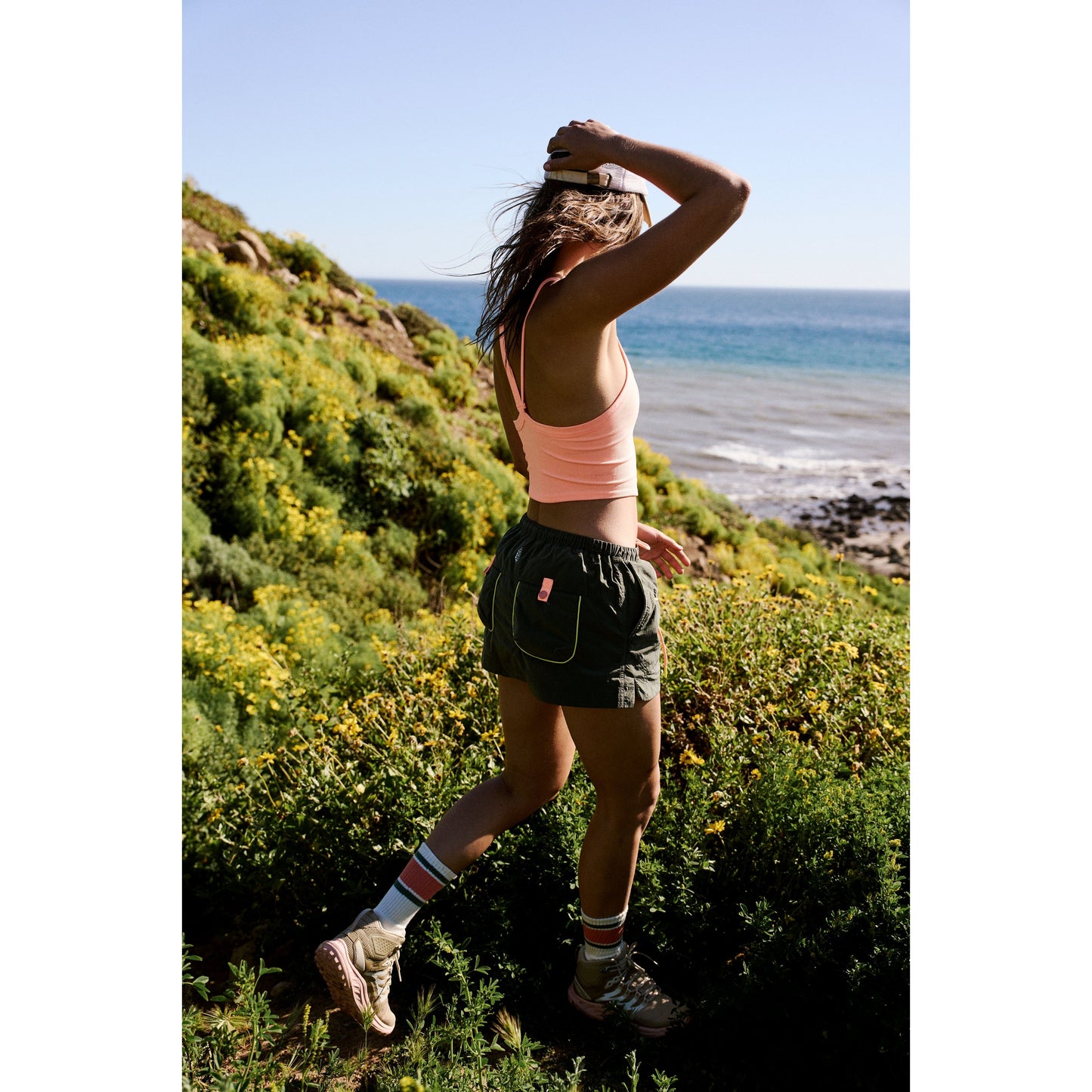 Woman jogging through coastal trail among wildflowers, sea in the background, tying her hair up in the Free People Movement Outskirts Skort in Olive Combo.