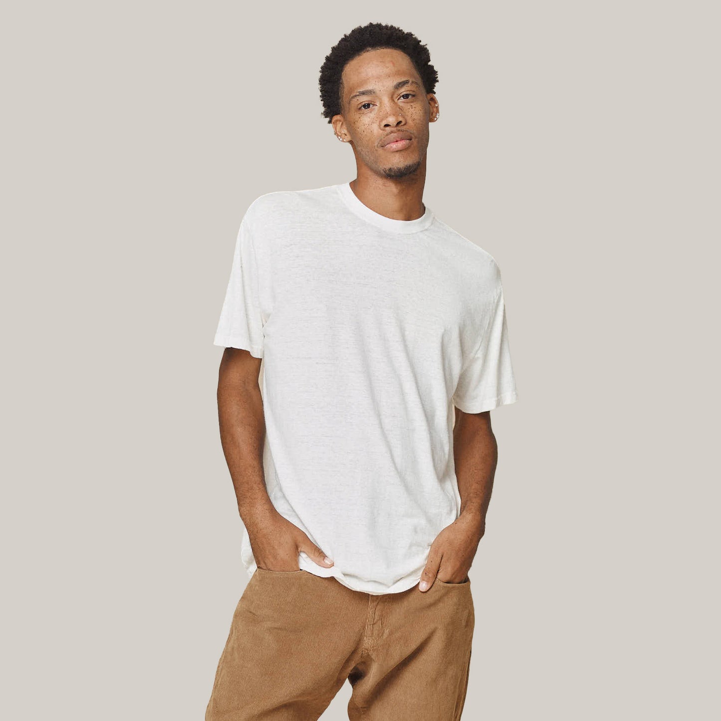 A man wears the Jungmaven Original Tee 5oz in Washed White with brown pants, standing with hands in pockets against a neutral background.
