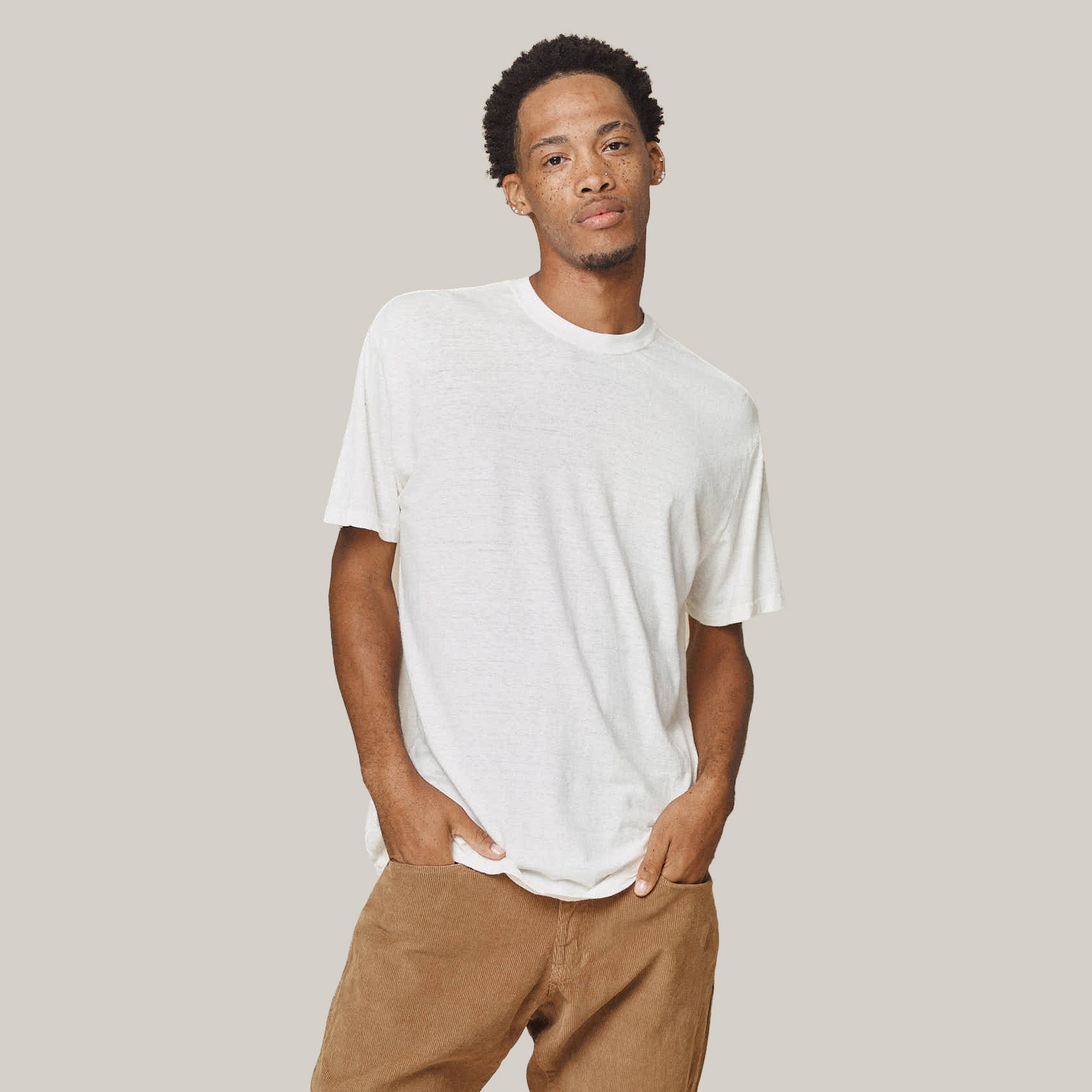 A man wears the Jungmaven Original Tee 5oz in Washed White with brown pants, standing with hands in pockets against a neutral background.