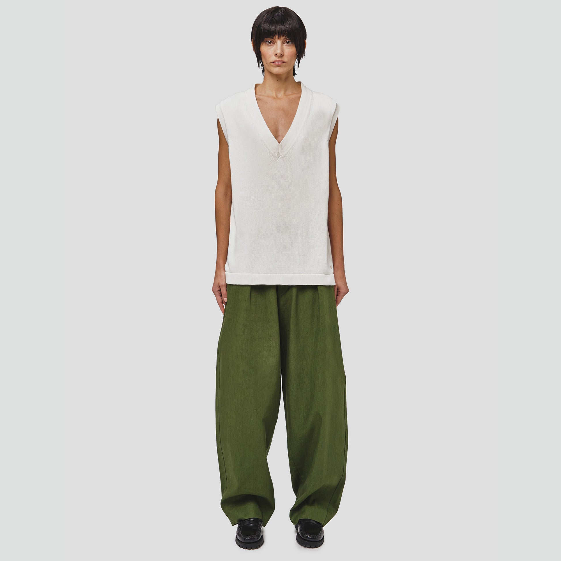 A model is seen wearing a Cotton Vest Sweater by Seven Gauge and loose green organic cotton pants, standing against a plain background.