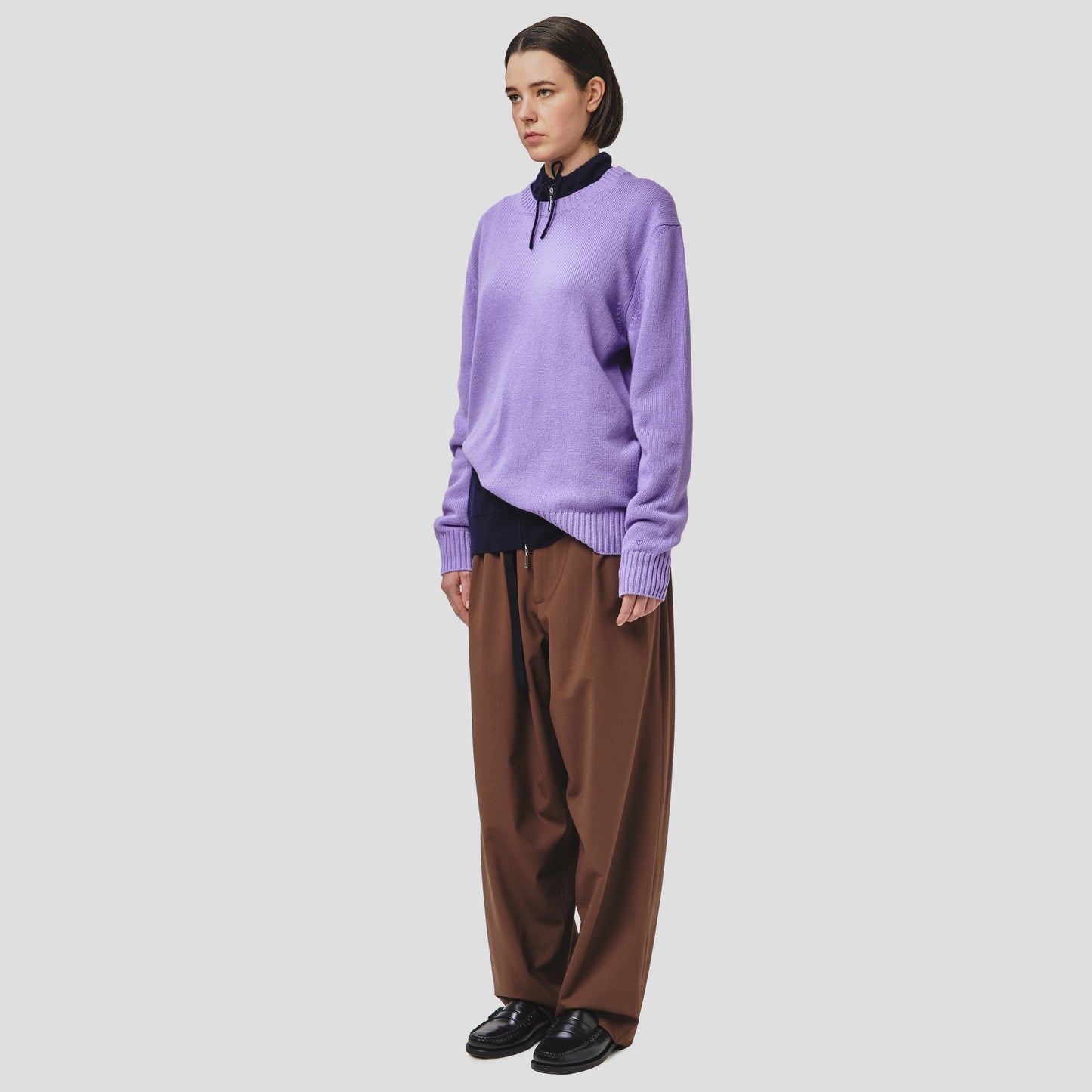 A person stands against a plain white background, wearing a Wool Cotton Crewneck Sweater by Seven Gauge in lilac, paired with a navy shirt, brown wide-leg pants, and black shoes.