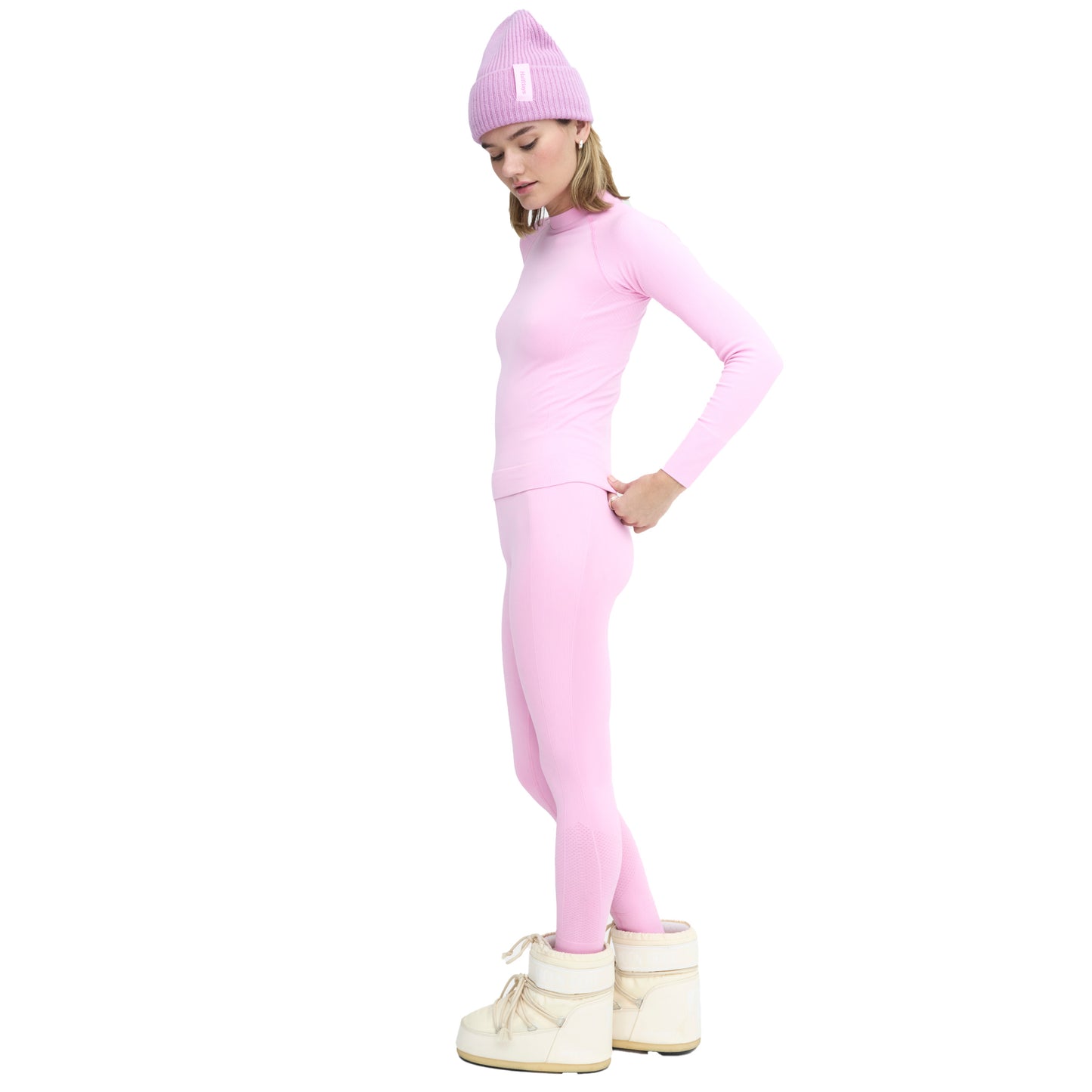 A person in a sleek pink outfit, showcasing the seamless Sophia Leggings by Halfdays and a beanie, stands in profile wearing white boots against a plain white background.