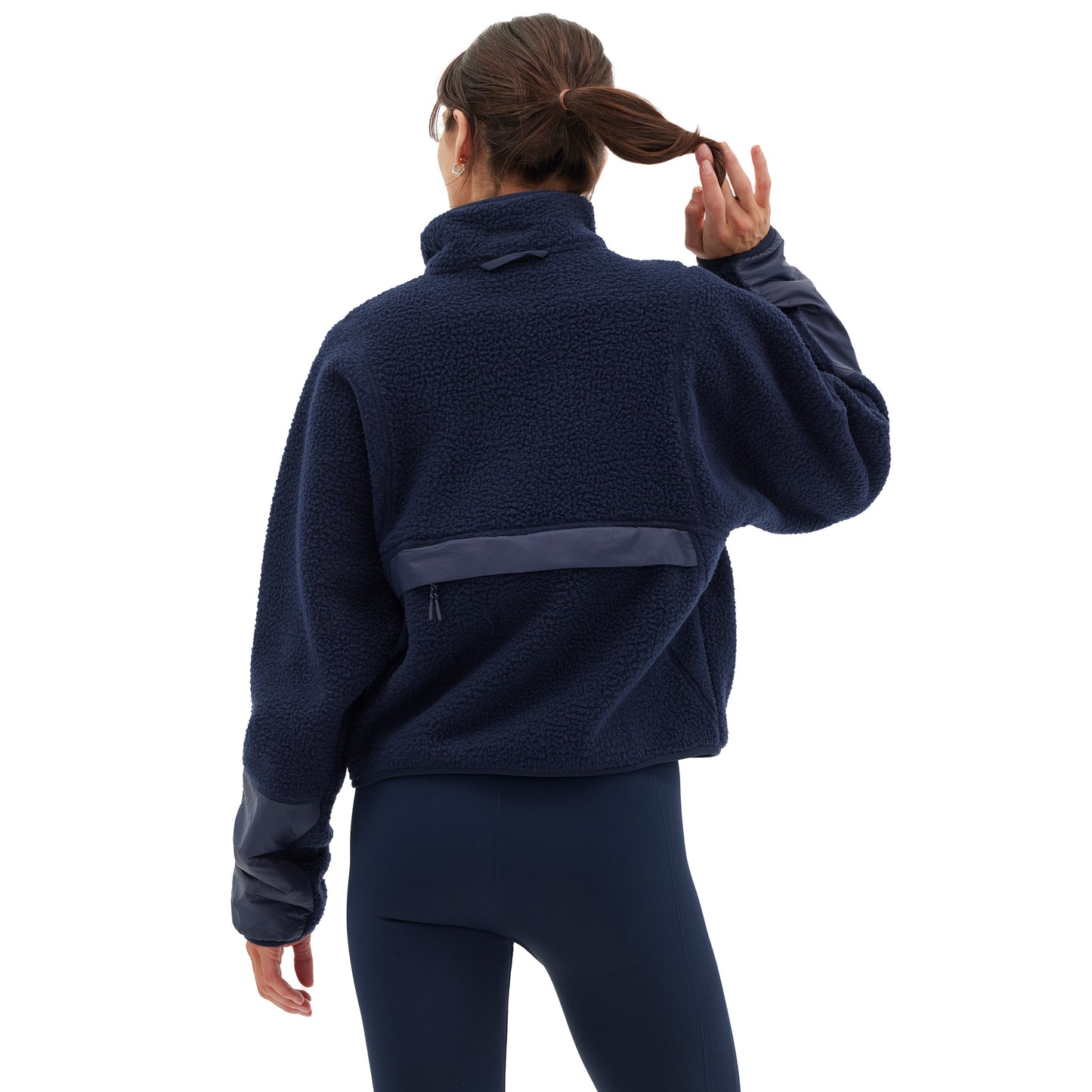 A person with a ponytail wearing the Halfdays Pieper Fleece in navy and dark leggings faces away, adjusting their hair, highlighting the jacket's performance durability.
