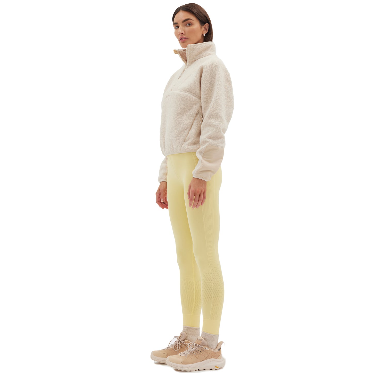 A woman in the Halfdays Pieper Fleece jacket and yellow leggings stands in profile against a white background, showcasing the blend of style and performance durability.