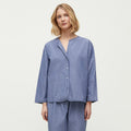 A woman stands barefoot, wearing a loose-fitting, long-sleeve pajama set made from Chetna Organic Cotton by Aiayu, featuring the Letto Casual Pant and a button-up top. She has shoulder-length blonde hair and a neutral expression. The background is plain white.