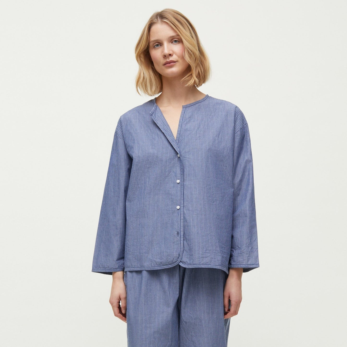 A woman stands barefoot, wearing a loose-fitting, long-sleeve pajama set made from Chetna Organic Cotton by Aiayu, featuring the Letto Casual Pant and a button-up top. She has shoulder-length blonde hair and a neutral expression. The background is plain white.