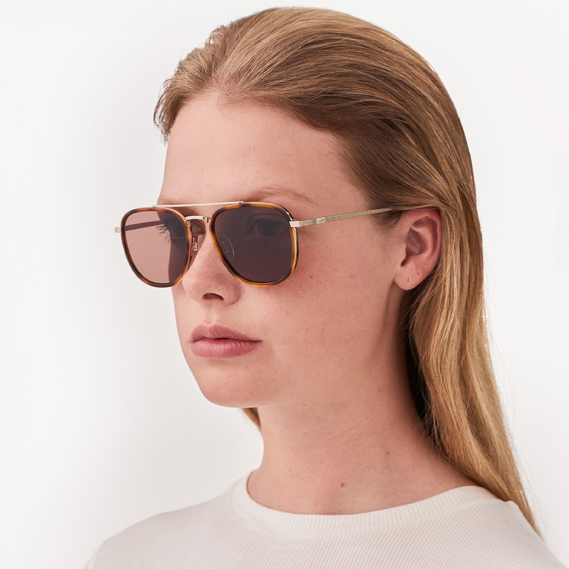 A person with long blonde hair wears Messyweekend Quentin sunglasses in Brown/Gold with high-quality lenses and a white top, set against a plain light background.