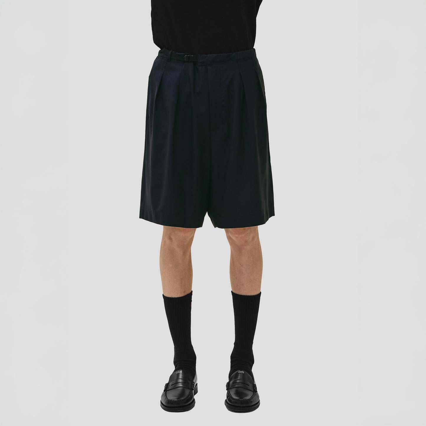 A person stands against a plain background wearing Seven Gauge's Tropical Wool Basket Short with an adjustable waist strap, along with black socks and shoes. Their upper body remains out of view.