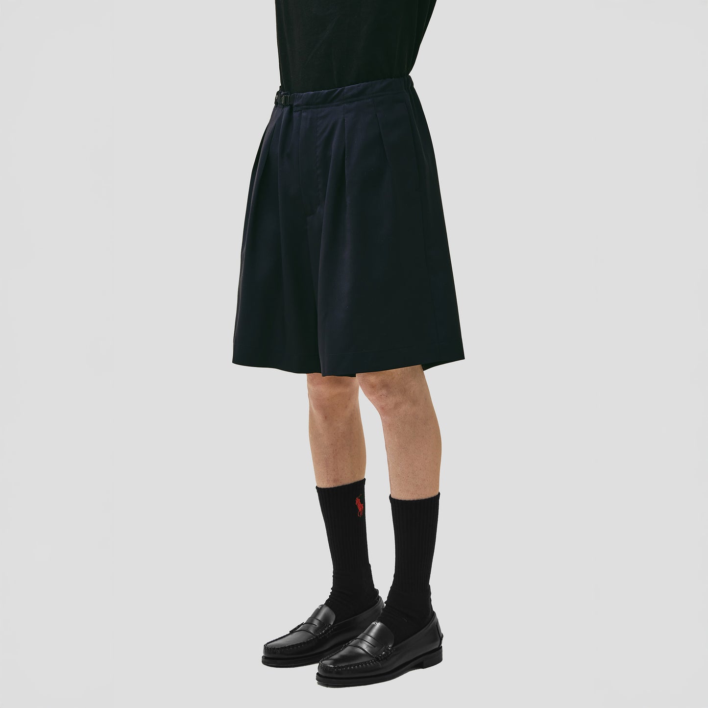 Someone is wearing Seven Gauge's Tropical Wool Basket Short in black with an adjustable waist strap, paired with black loafers and socks, against a plain white background.
