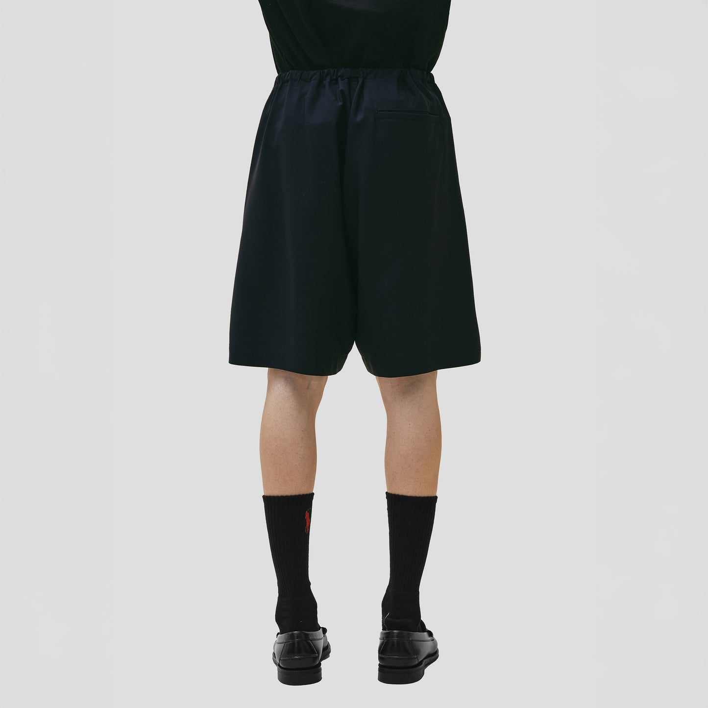 A person in Seven Gauge's Tropical Wool Basket Short with adjustable waist, black socks, and comfy fit black shoes stands against a white background.