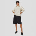 A person stands against a plain background wearing a beige hoodie, Seven Gauge's Tropical Wool Basket Short with an adjustable waist, and black loafers, with their hands in the hoodie pocket.