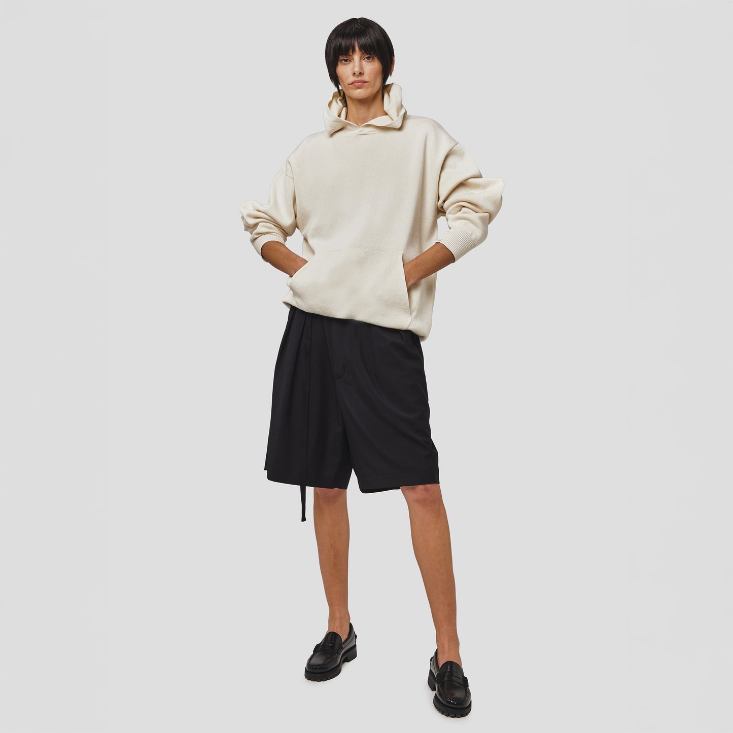 A person stands against a plain background wearing a beige hoodie, Seven Gauge's Tropical Wool Basket Short with an adjustable waist, and black loafers, with their hands in the hoodie pocket.
