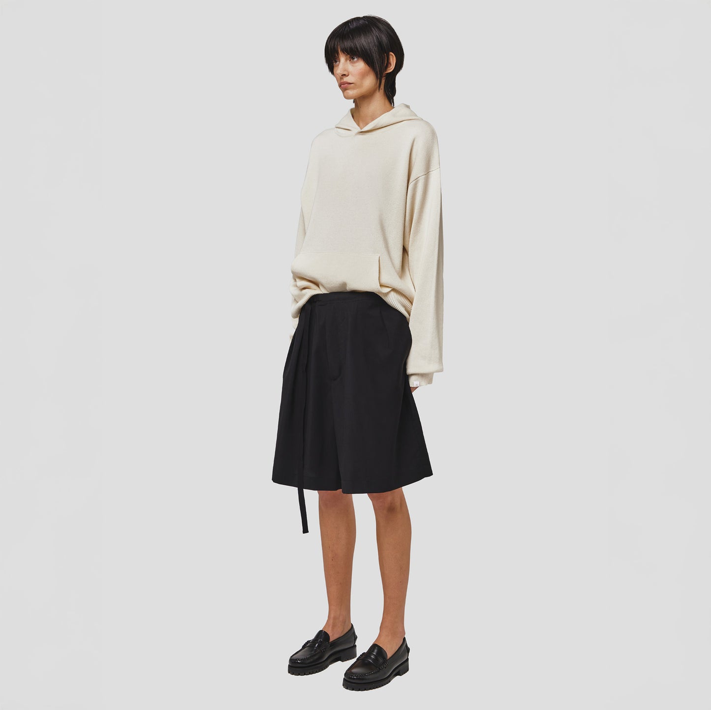 The person wears a beige hoodie, a Tropical Wool Basket Short by Seven Gauge, and black loafers while standing against a plain white background, exuding a comfort fit.