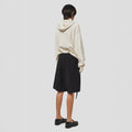 A person wearing a cream hoodie and black Tropical Wool Basket Shorts by Seven Gauge, featuring an adjustable waist strap, stands facing away against a plain background.