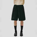 A person stands against a plain gray background, wearing Seven Gauge's Tropical Wool Basket Short with an adjustable waist strap for comfort, complemented by a cream shirt, black socks, and shoes.