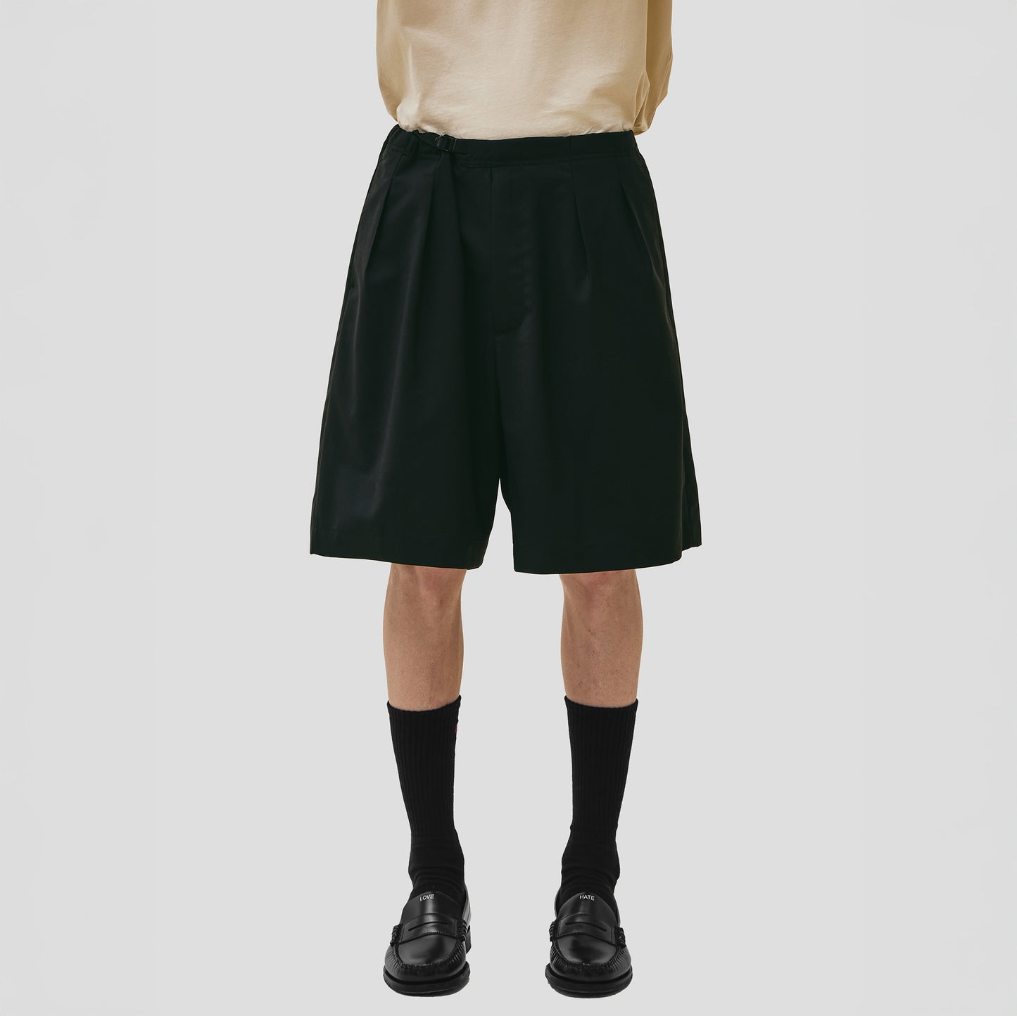 A person stands against a plain gray background, wearing Seven Gauge's Tropical Wool Basket Short with an adjustable waist strap for comfort, complemented by a cream shirt, black socks, and shoes.