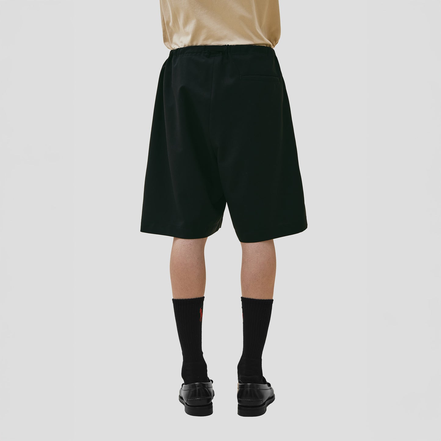 Rear view of a person wearing a beige shirt and Seven Gauge's Tropical Wool Basket Short with an adjustable waist strap, paired with black socks and shoes against a plain background.