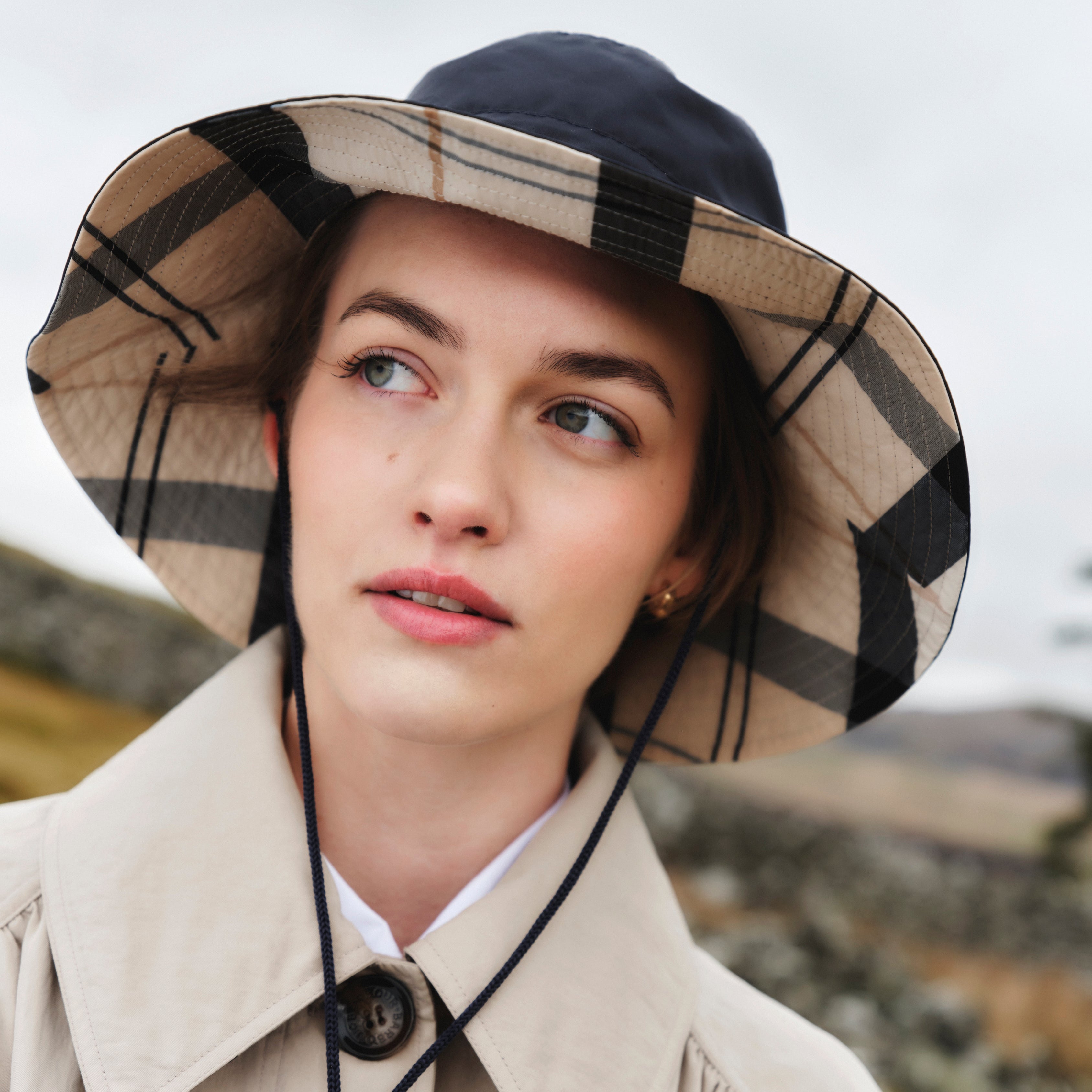 A woman in a Barbour Darla Showerproof Bucket Hat and beige trench coat looks to the side outdoors on a cloudy day.