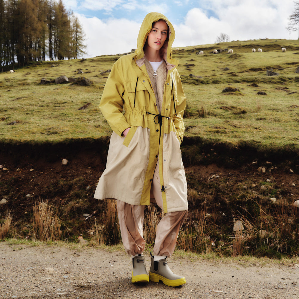 A person in a Barbour Evie Showerproof Parka with an adjustable hood, beige pants, and boots stands on a dirt path in a grassy field. Scattered sheep roam among the trees, enjoying superior weather protection.