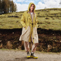 A person in a Barbour Evie Showerproof Parka with an adjustable hood, beige pants, and boots stands on a dirt path in a grassy field. Scattered sheep roam among the trees, enjoying superior weather protection.