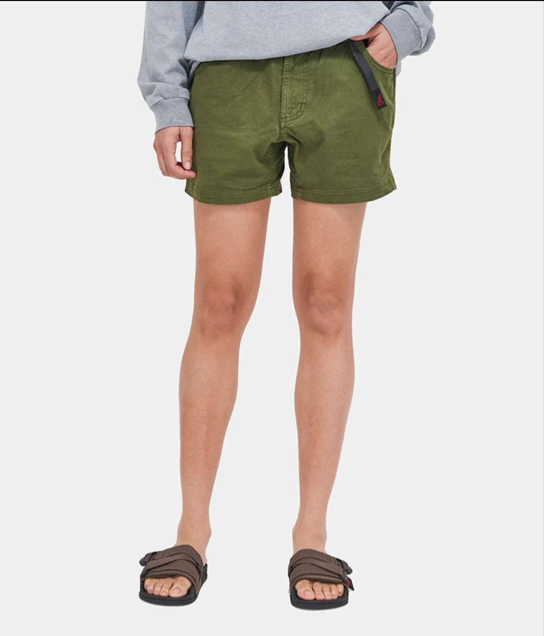A person stands against a plain white background wearing an untucked grey top, brown sandals, and Westerlind Very Short in Olive.