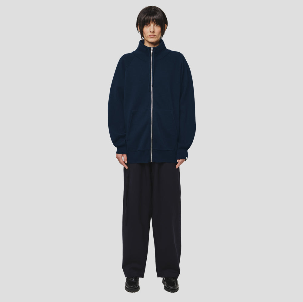 A person with short dark hair and a neutral expression stands against a light gray background, wearing the Seven Gauge Wool Cotton Full Zip Sweater in Blu, wide-leg pants, and black shoes.