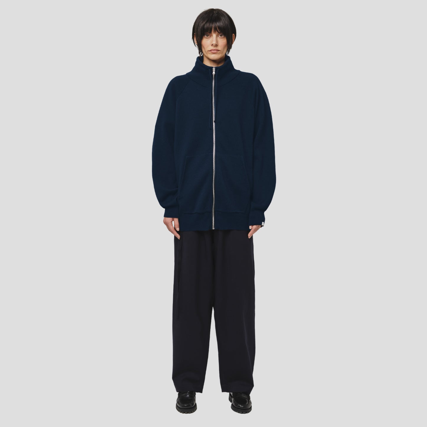 A person with short dark hair and a neutral expression stands against a light gray background, wearing the Seven Gauge Wool Cotton Full Zip Sweater in Blu, wide-leg pants, and black shoes.