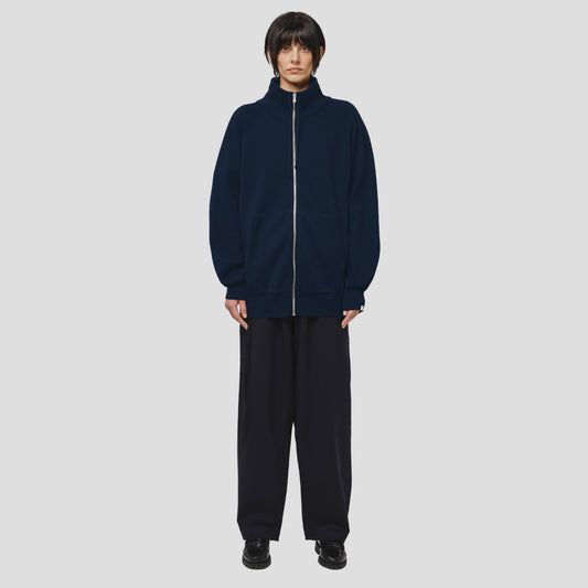 A person with short dark hair and a neutral expression stands against a light gray background, wearing the Seven Gauge Wool Cotton Full Zip Sweater in Blu, wide-leg pants, and black shoes.