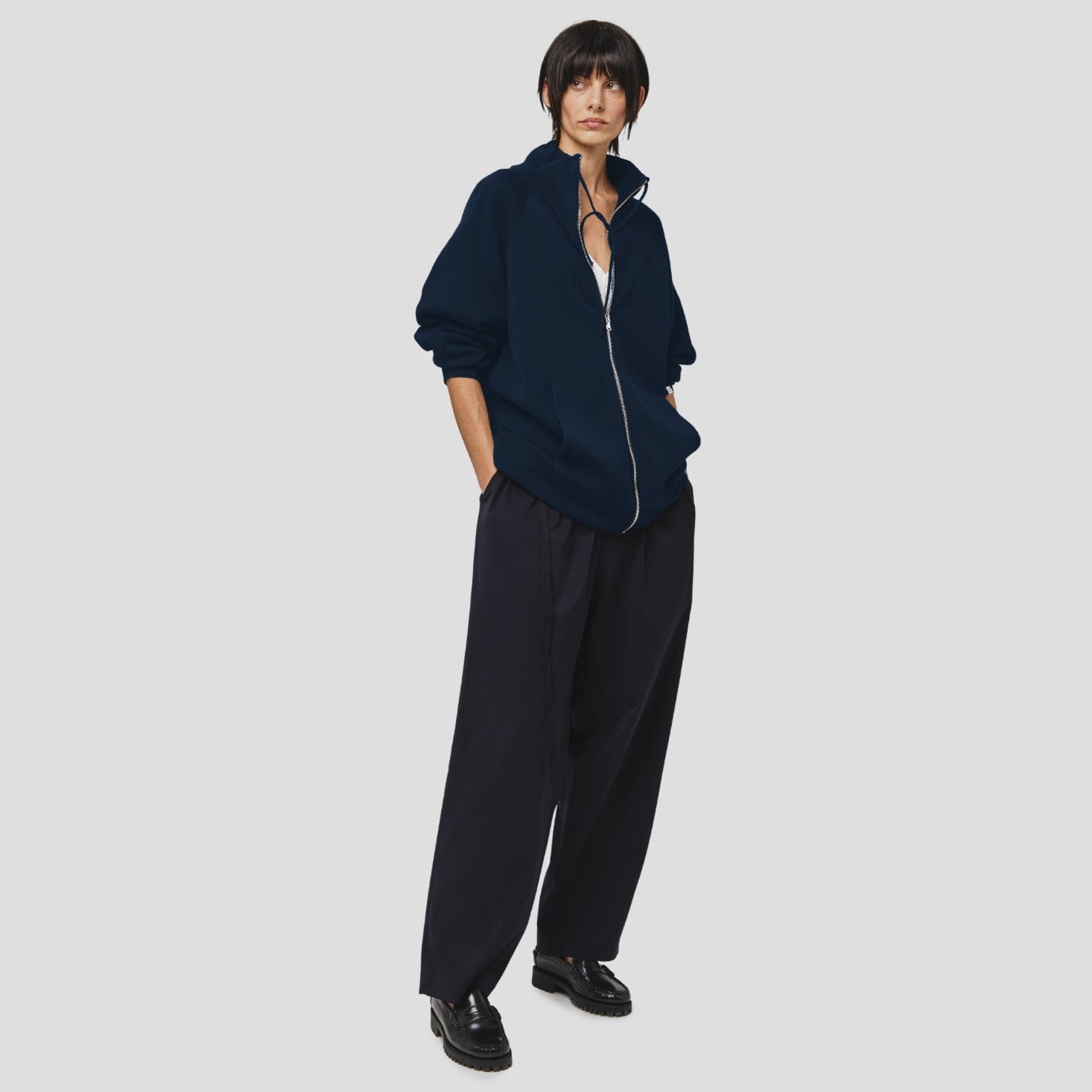 A person stands posing in a Seven Gauge Wool Cotton Full Zip Sweater in Blu, paired with loose dark pants and black shoes, against a plain light gray background.