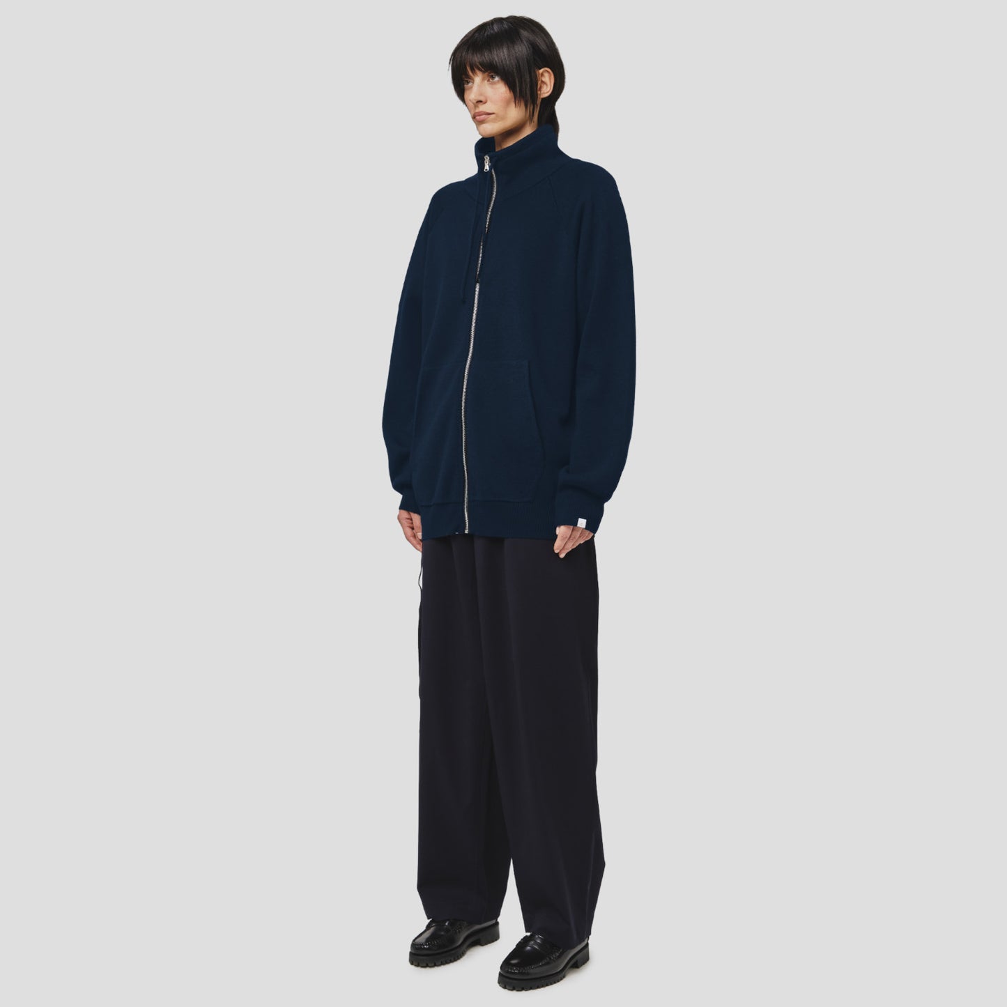 Person standing against a plain background, wearing the Seven Gauge Wool Cotton Full Zip Sweater in Blu, wide-leg pants, and black shoes, with hands by their sides.
