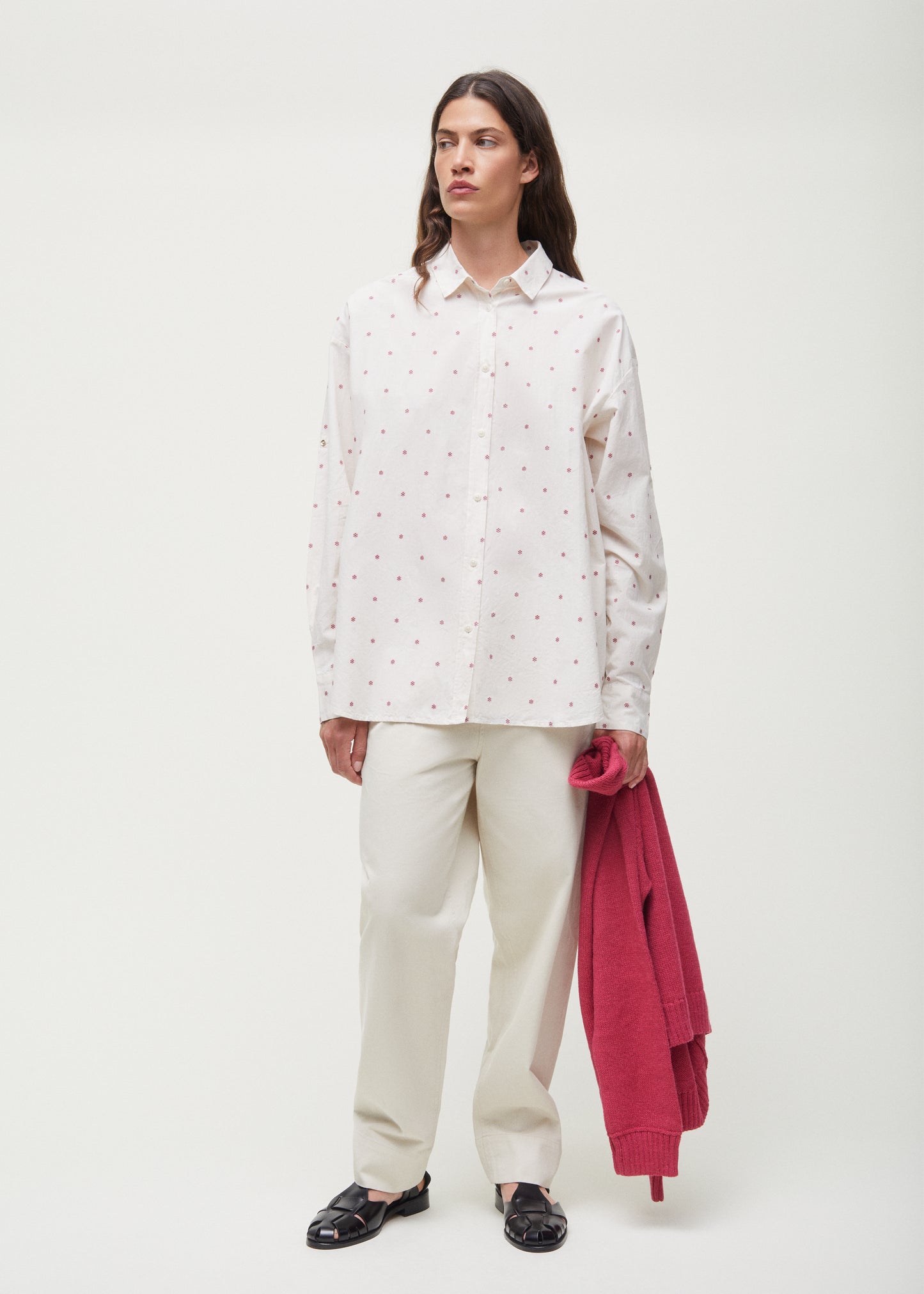 An individual dressed in a white, patterned Aiayu Shirt Flore crafted from organic cotton and light-colored pants, while holding a red sweater.