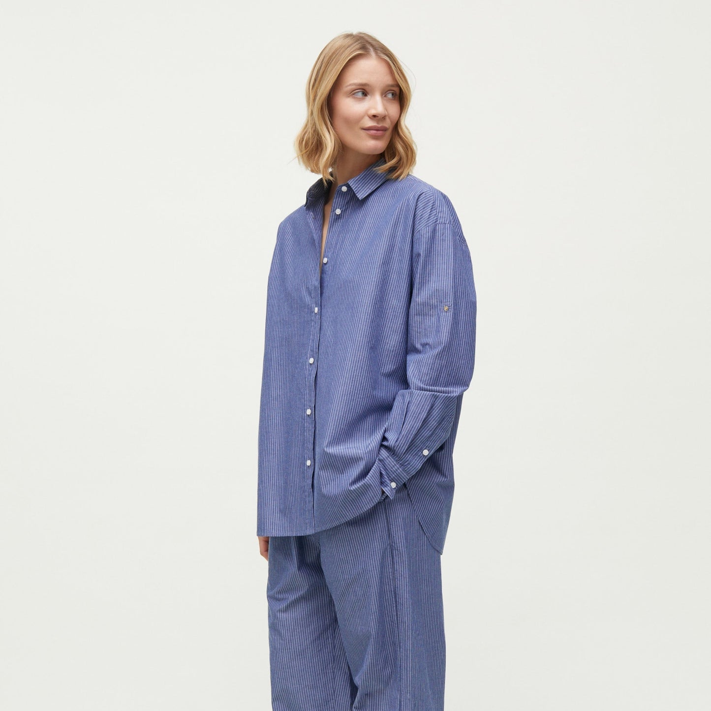 A person stands wearing Aiayu's Letto Casual Pant and a loose-fitting blue button-up shirt, both made from Chetna Cotton sourced from India, with light-colored socks. They are looking slightly to the left and smiling against a plain white background.
