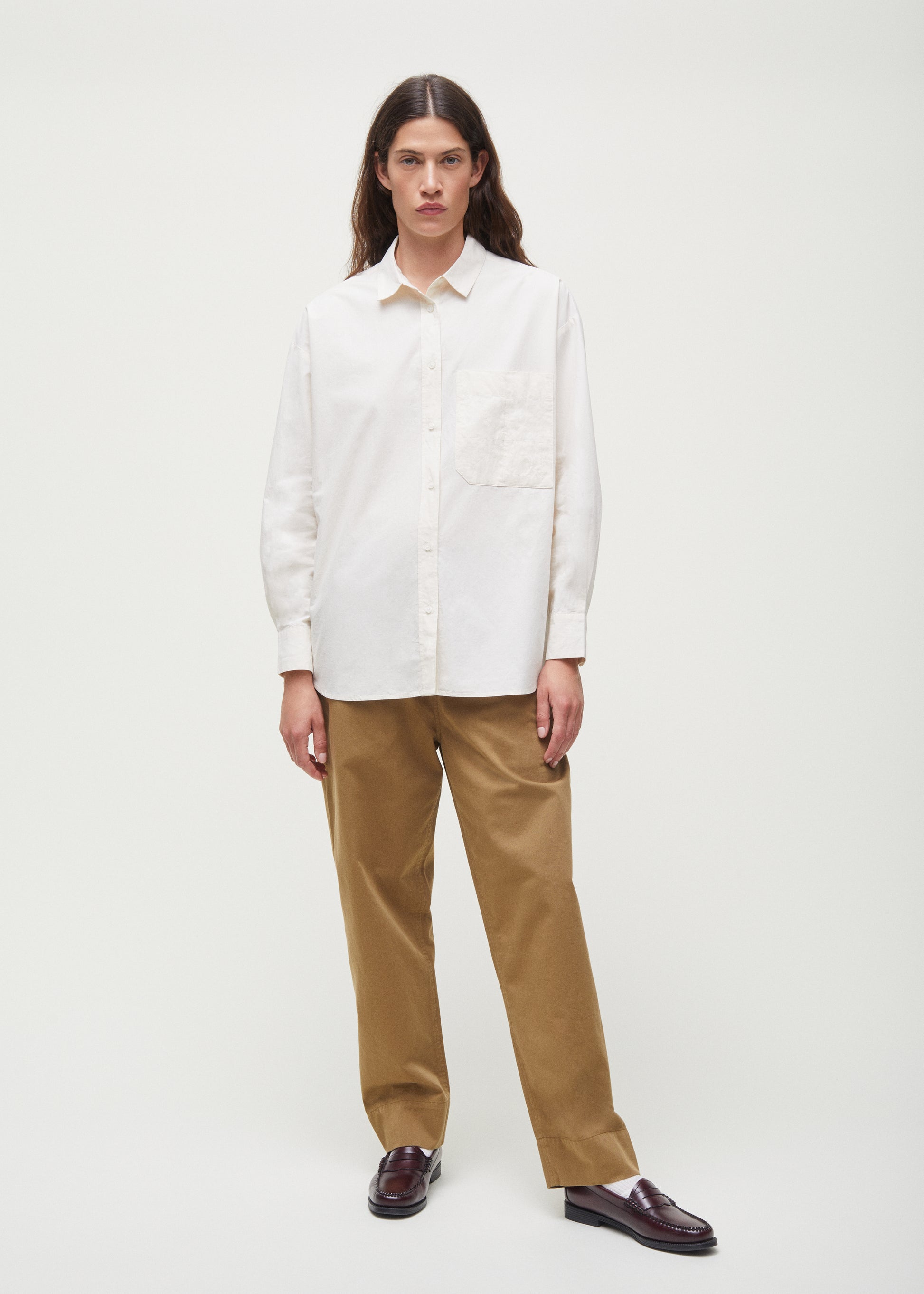 A person with long hair is wearing Aiayu's oversized-fit Shirt Quilt, made from regenerative cotton, along with khaki pants and dark loafers, standing against a plain background.
