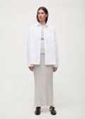 A person stands against a plain background wearing a white button-up shirt over the Seinne Linen Skirt by Aiayu, made from European organic cotton, paired with black shoes. Their hands rest at their sides.