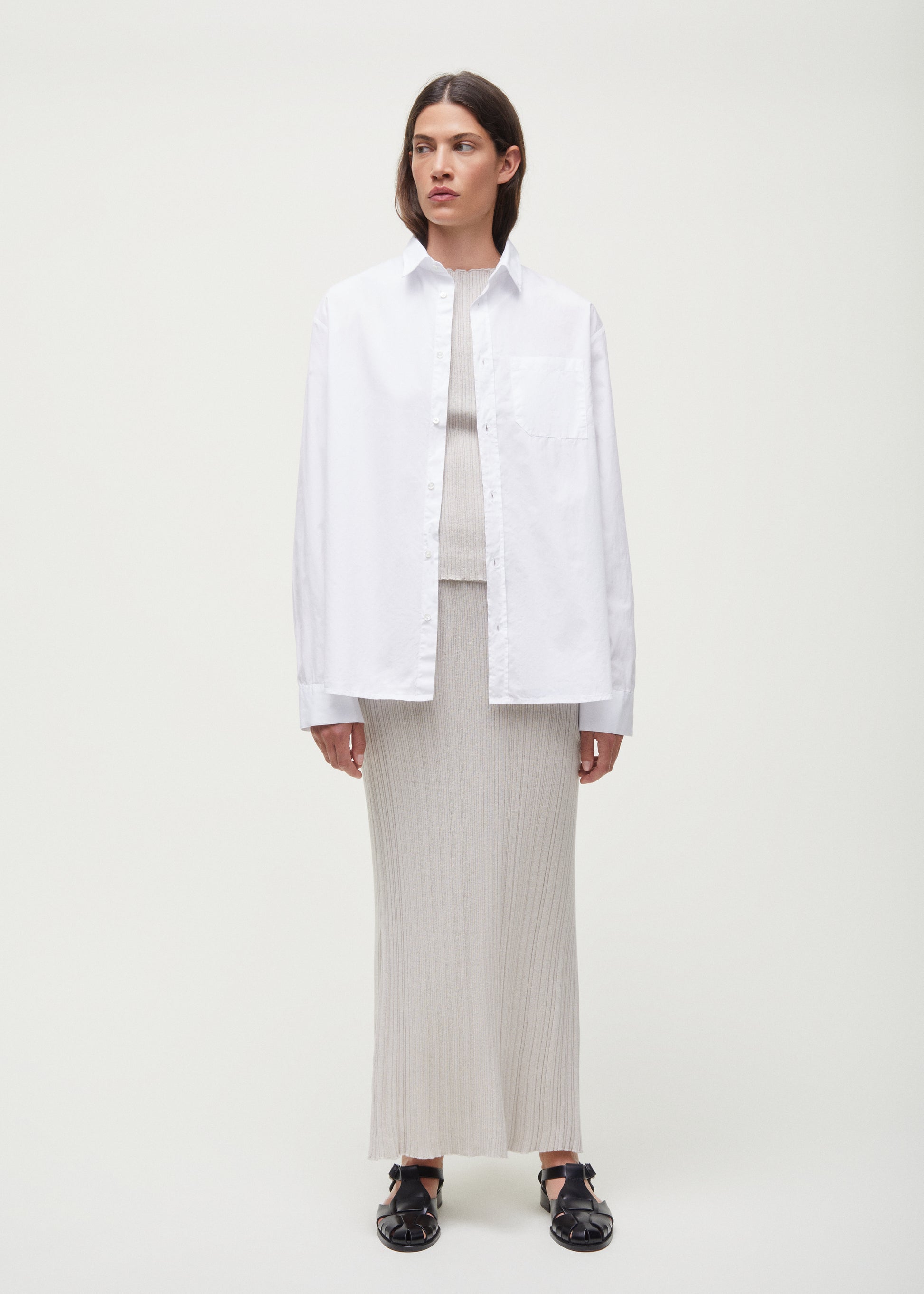 A person stands against a plain background wearing a white button-up shirt over the Seinne Linen Skirt by Aiayu, made from European organic cotton, paired with black shoes. Their hands rest at their sides.