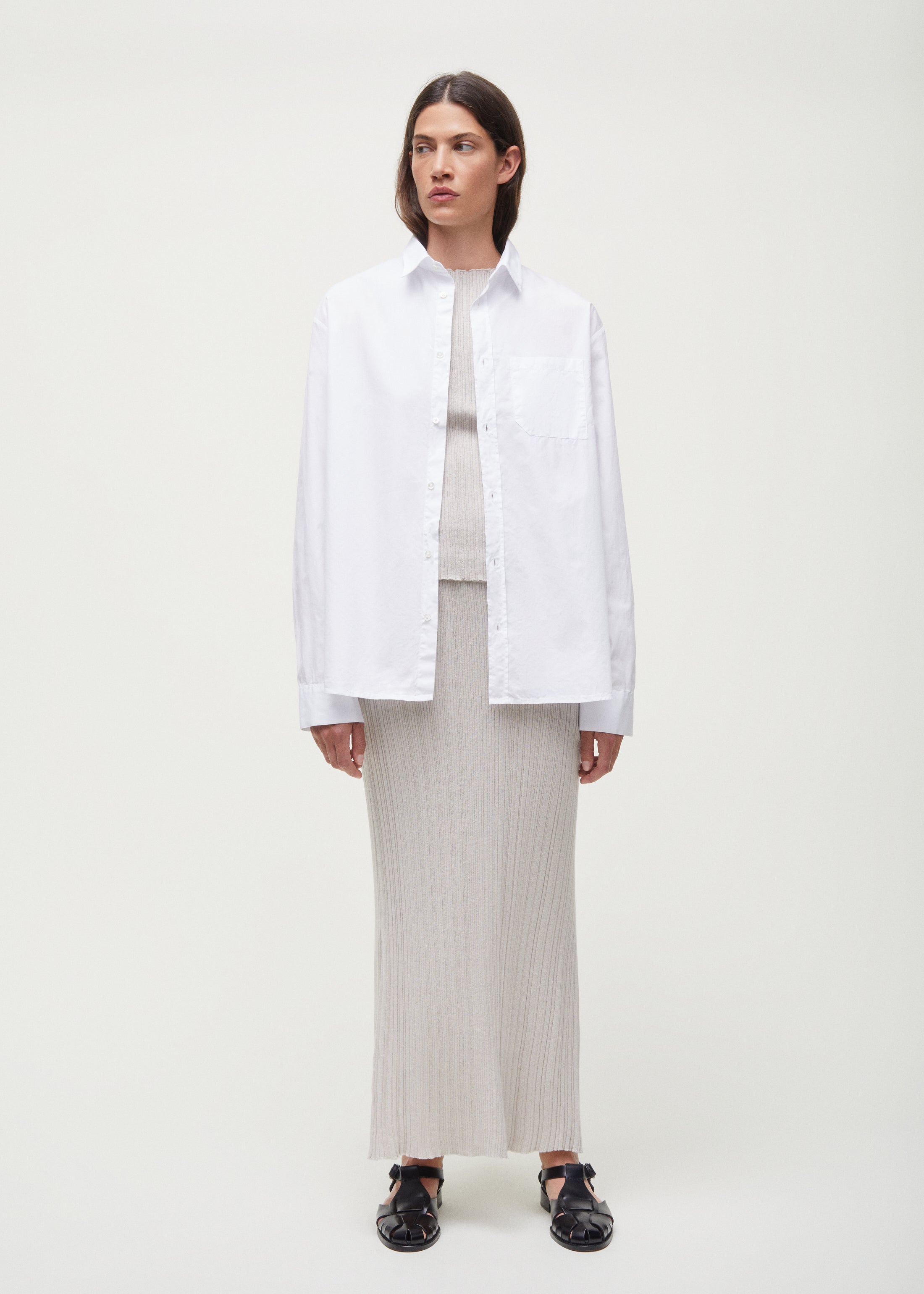 A person stands against a plain background wearing a white button-up shirt over the Seinne Linen Skirt by Aiayu, made from European organic cotton, paired with black shoes. Their hands rest at their sides.