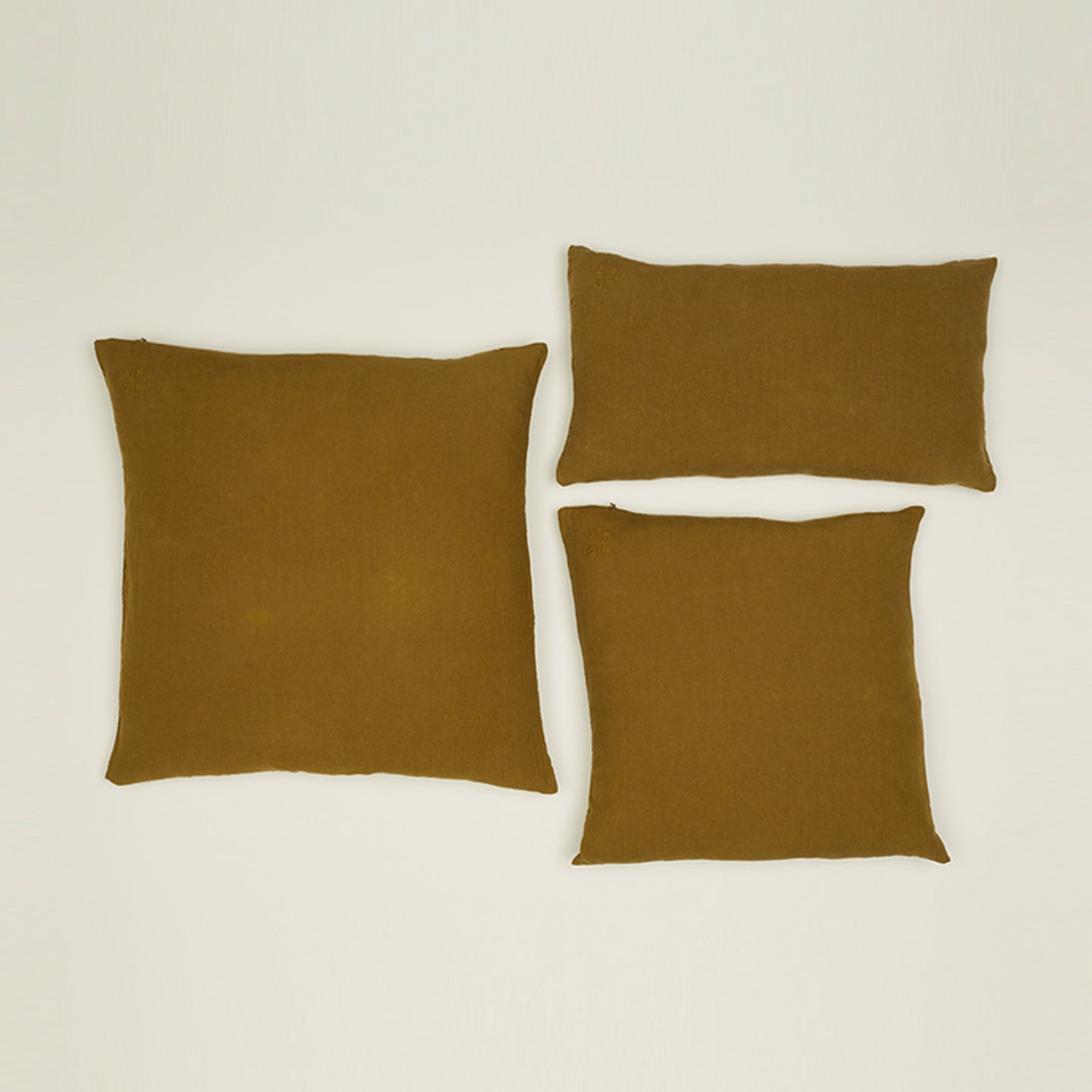 Three cushions on a light surface; two square "Simple Linen Pillows - 18x18, Bronze" by Hawkins New York and one rectangular, made from soft European linen.