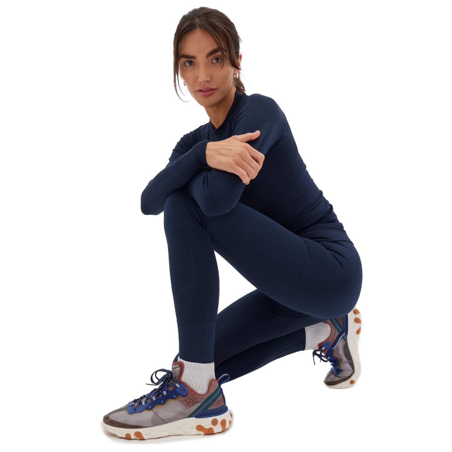 A person dressed in navy athletic wear, including the Sophia Legging by Halfdays, is squatting on one knee. Complementing their adventure-ready look are white socks and colorful sneakers, all while they maintain a neutral expression.