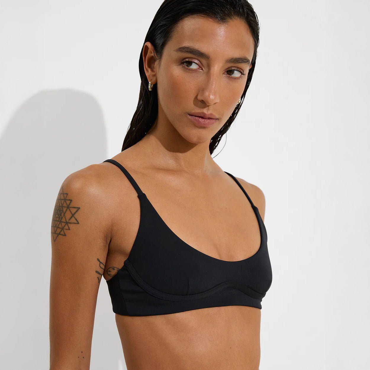 A person with long dark hair wears the Left On Friday Club Top bralette, standing against a white background and looking to the side. A geometric tattoo is visible on their upper arm.