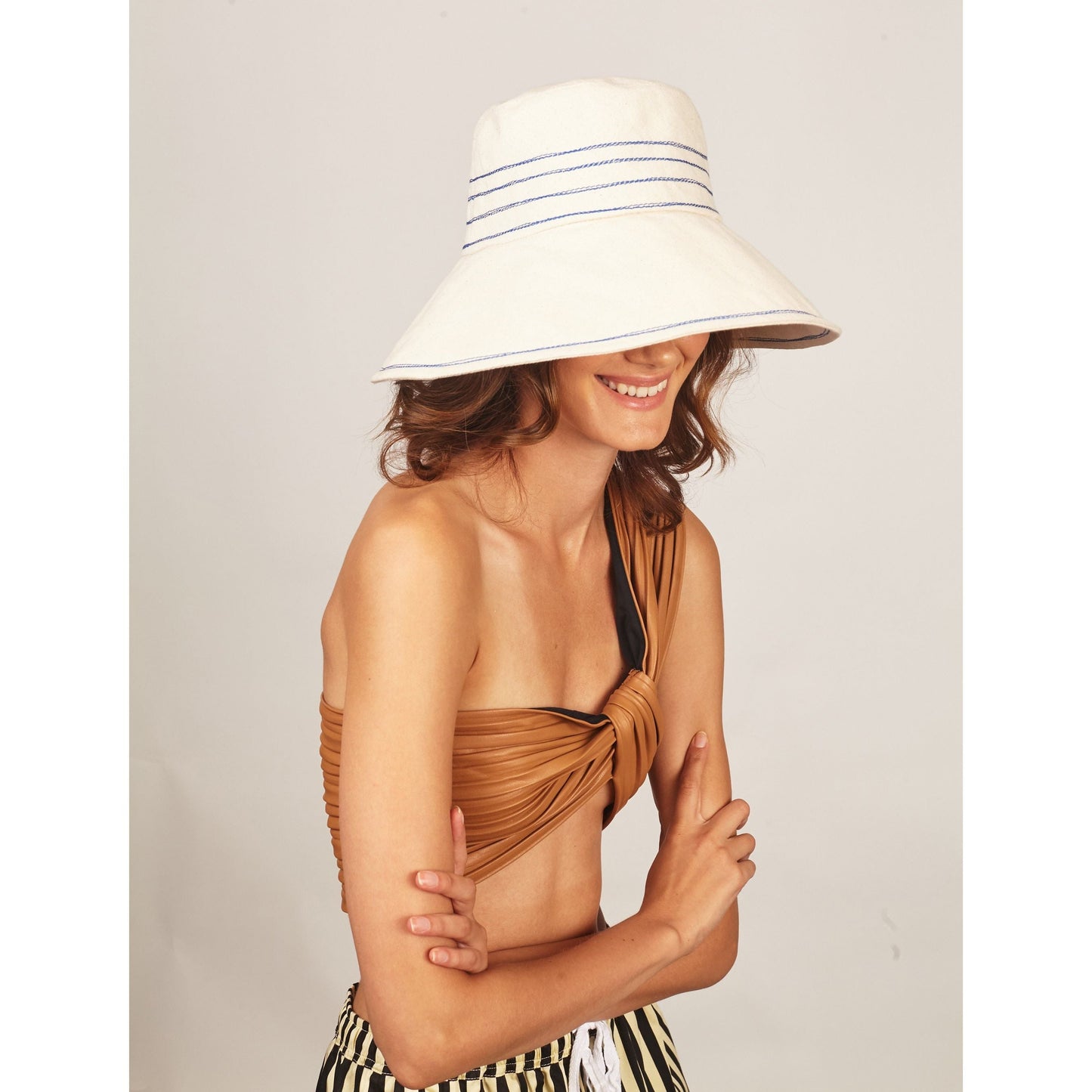 Woman smiling, wearing a Tide Lines hat by Lola Hats with cobalt blue stitching, a brown twisted bandeau top, and black and white striped pants.