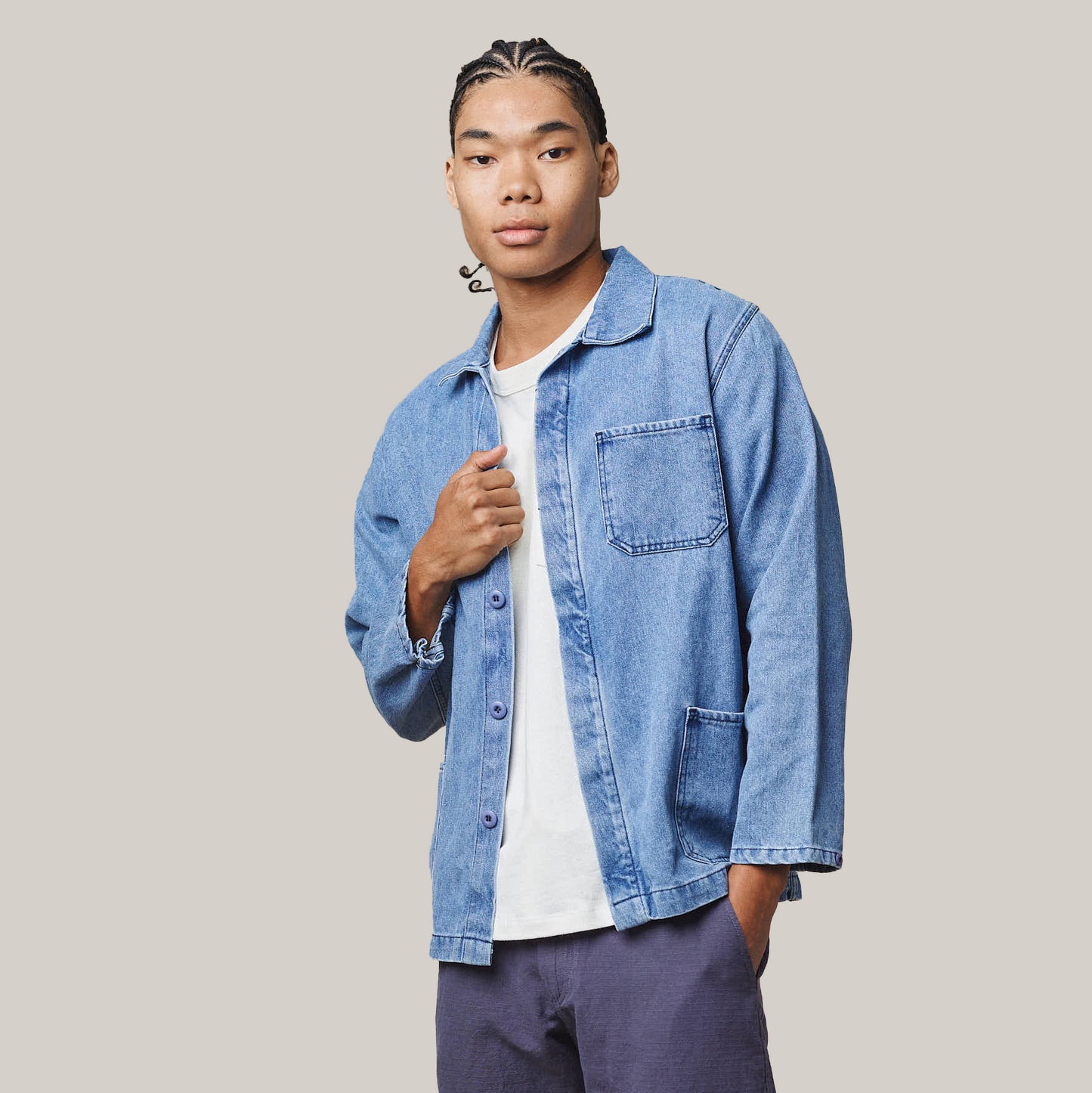 A person with braided hair wears the Jungmaven Olympic Jacket in Light Denim over a white shirt and purple pants, standing against a plain, light background.