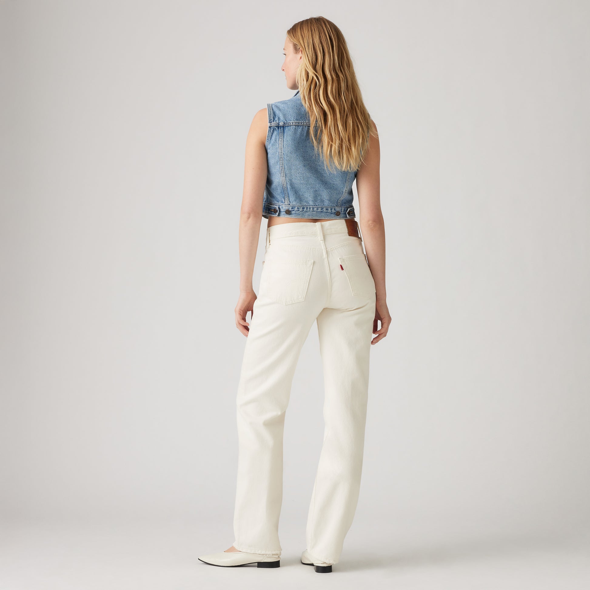 A woman faces away, wearing a sleeveless denim vest, Levis 501 90s mid-rise white jeans, and white shoes against a plain light background.