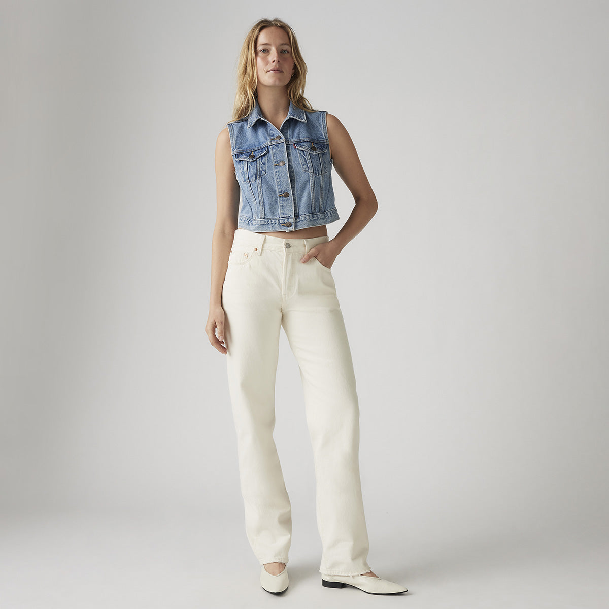 A woman poses against a plain background wearing a sleeveless denim vest, Levi’s 501 90s vintage blue jeans, and white shoes.